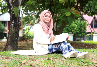 Portrait of smiling young woman reading book while sitting at park