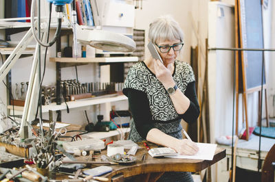 Senior woman using mobile while writing on document in jewelry workshop