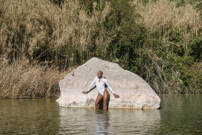 Self assured stylish female with short hair wearing trendy swimsuit standing near rock in rippling water in sunny weather