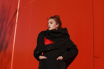 Fashionable teenage girl wearing black warm clothing standing against red wall during sunny day