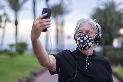 Senior man wearing mask taking selfie with smart phone in park