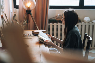 Young smiling woman teenager girl student with dark long hair in casual reading book at the home