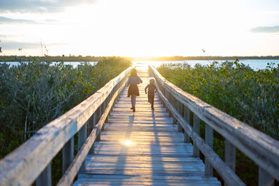 Rear view of siblings running on pier leading towards sea against cloudy sky