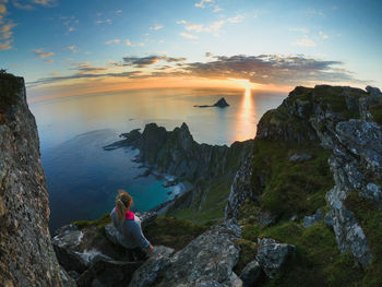 Rear view of woman sitting on rock against sky during sunset