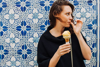 Millennial woman licking finger while eating ice cream at a tiled wall