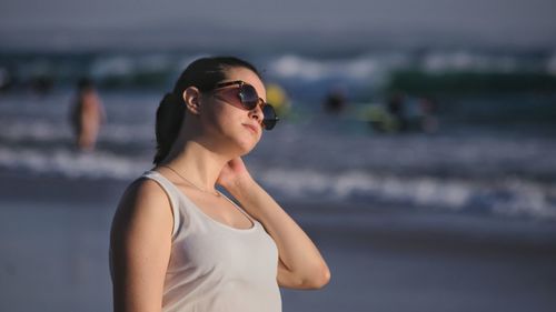 Young woman wearing sunglasses standing at beach against sky