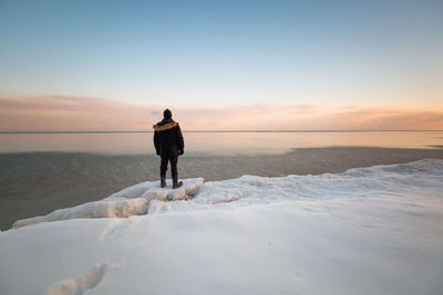 Rear view of man looking at sea while standing on snow during sunset