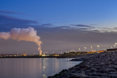 Illuminated factory by sea against sky