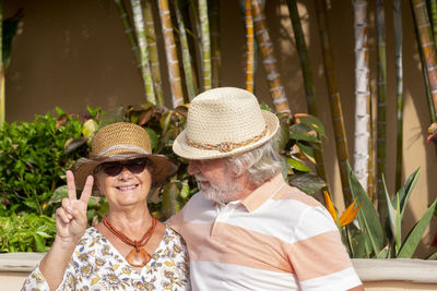 Portrait of senior couple showing symbol of peace wearing hat against plants