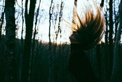 Woman tossing hair while standing against trees during sunset