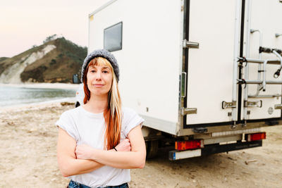 Positive female traveler standing with crossed arms near truck parked at lakeside in mountains and looking at camera