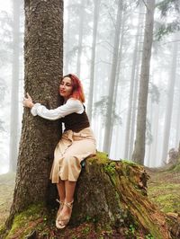 Full length portrait of woman standing by tree trunk in forest