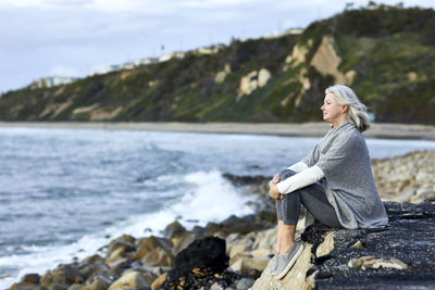 Side view of senior woman looking at sea while sitting on rock during sunset