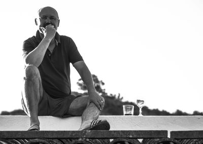 Low angle portrait of mid adult man having drink while sitting on building terrace against clear sky