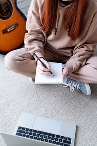 Young caucasian red-haired woman learning to play acoustic guitar at home, making notes 