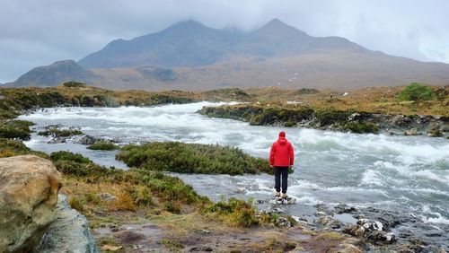 Rear view of person standing by flowing river against mountains