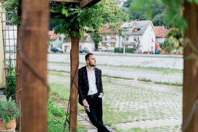 Young man holding camera and looking away while standing by wooden post