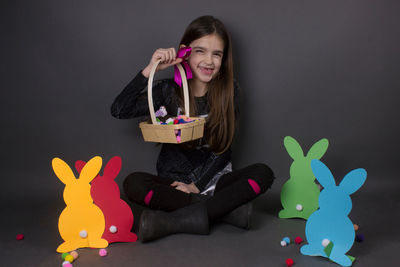 Portrait of smiling girl with eater bunny and pom pom in basket against gray background