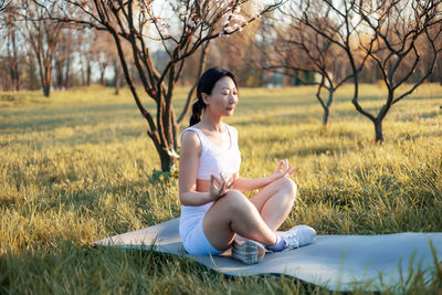 Asian woman practicing yoga sitting on grass, meditating with closed eyes under the tree.