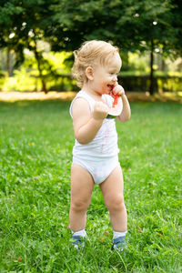 Funny smiling kid boy in white bodysuit eating watermelon at green lawn