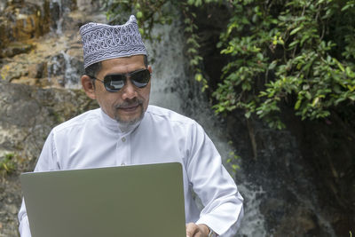 Mature man wearing sunglasses while using laptop against waterfall