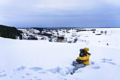 Young cheerful woman in yellow sitting in snow holding fluffy gray dog, traveling with pets in w