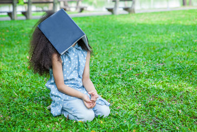 Bored girl covering face with book on grassy land in park