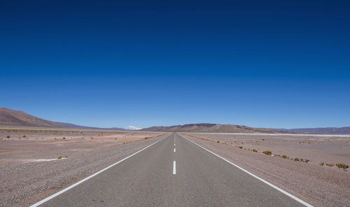 Road leading towards desert against clear blue sky