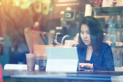 Woman using digital tablet by coffee cup while sitting on table at cafe seen through glass window