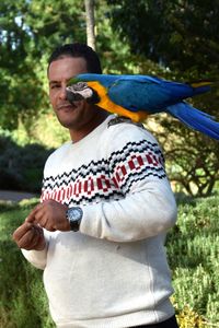 Portrait of man with macaw on shoulder standing against trees