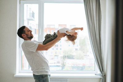 Young man father having fun with baby girl near window at home