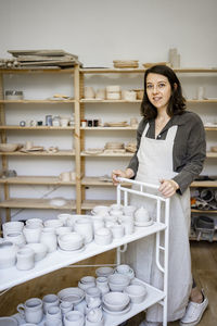 Smiling young craftswoman with pottery cart in workshop