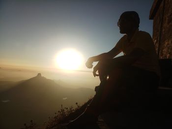 Silhouette man sitting on mountain against sky during sunset
