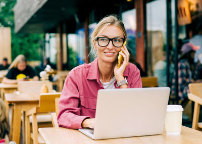 Young businesswoman talking on the phone and using a laptop while sitting in a cafe