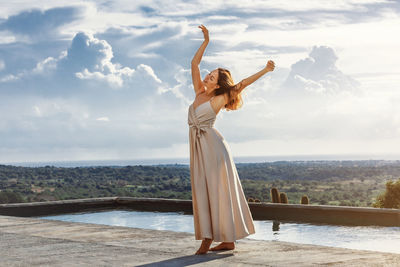 Young brunette woman dancing next to a pool, wearing a cream white dress with a moody sky