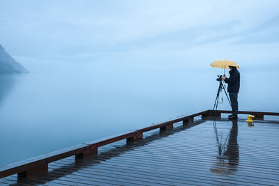 Full length of man standing on pier with tripod and umbrella during monsoon