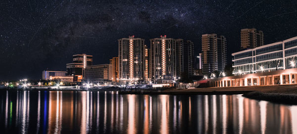 Illuminated modern buildings by sea against sky at night