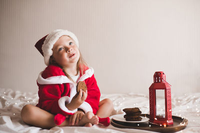 Cute child girl 3-4 year old wear red santa claus hat and bathrobe sit in bed with biscuit 