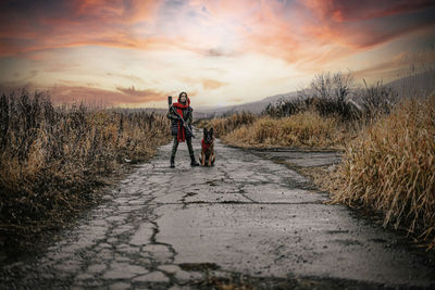 Full length of woman holding gun walking on dirt road against sky