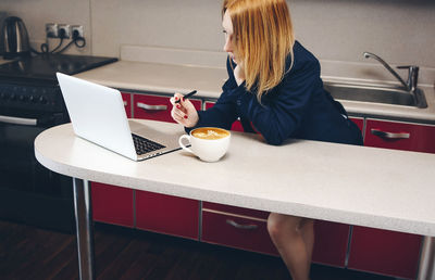 Woman working on laptop while leaning on table in kitchen