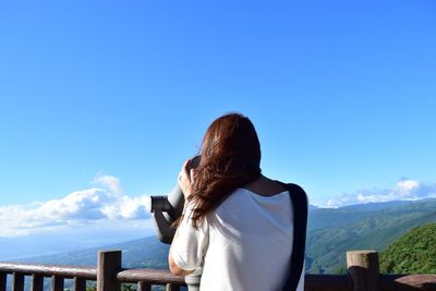 Rear view of woman looking through binoculars against blue sky