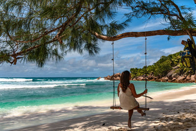 Rear view of young woman wearing beach clothes, sitting on swing on paradise beach