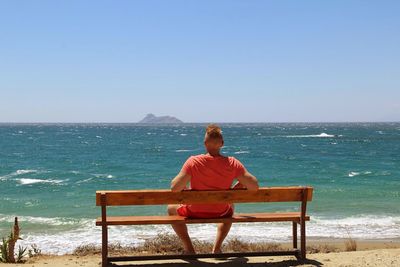 Rear view of man sitting on bench at beach against clear sky