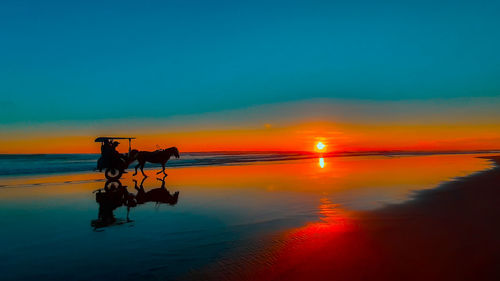 A silhouetted horse-drawn carriage at sunset time on the wet and reflecting sand beach on java.