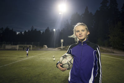 Portrait of girl standing with soccer ball on field against trees at night