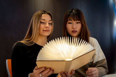 Multi-ethnic female couple of girlfriends are looking at a magical bright book indoors