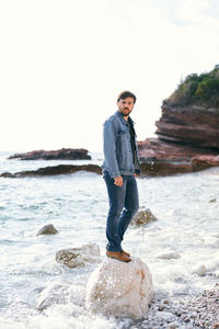 Young man standing on rock at sea shore against sky