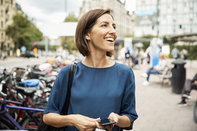 Smiling mid adult businesswoman looking away while holding smart phone in city