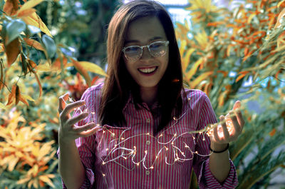 Happy woman holding illuminated string lights amidst plants