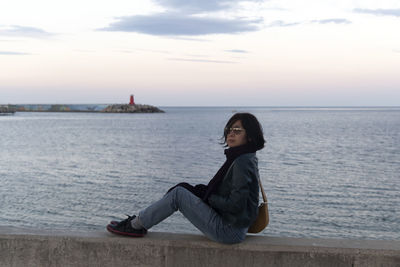 Side view of woman sitting on rock by sea against sky